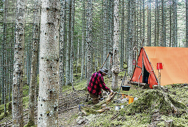 Senior man preparing campfire to cook food near tent in forest