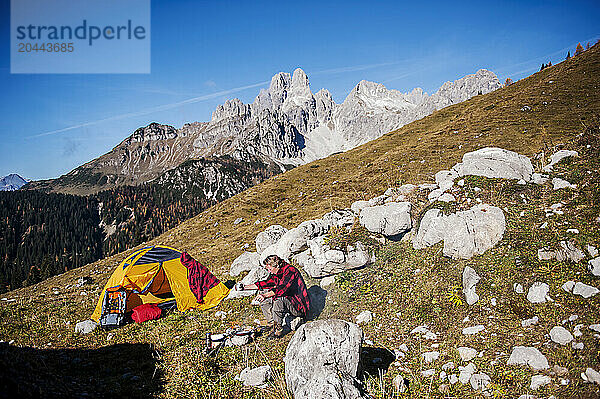 Senior man cooking food near rocks and camping on mountain