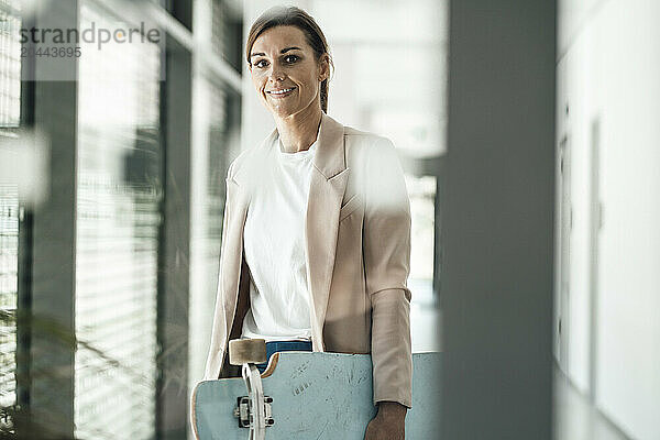 Happy businesswoman holding skateboard and standing at office