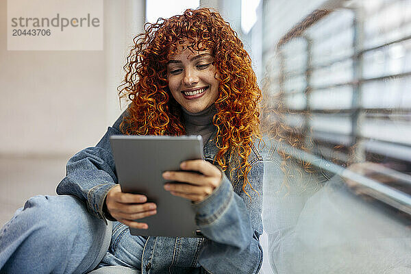 Smiling redhead young woman with curly hair using tablet PC leaning on window glass