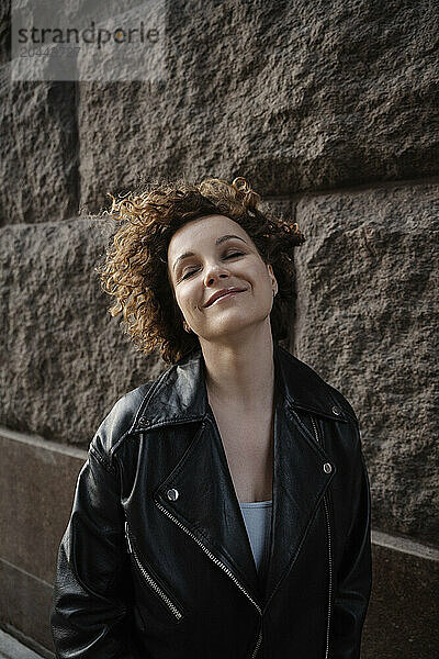 Smiling woman with eyes closed standing near wall