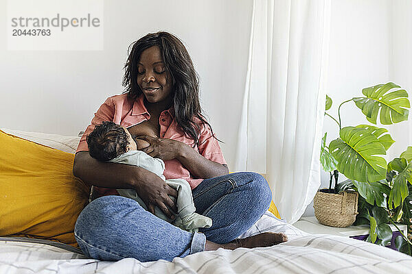 Mother breastfeeding daughter and sitting on bed at home