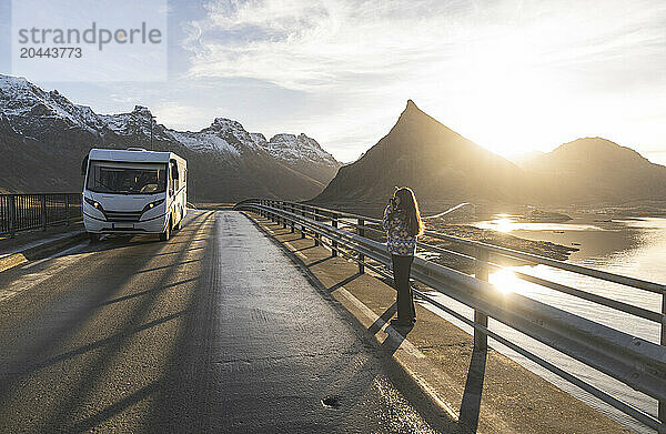 Norway  Nordland County  Young woman photographing motor home standing on coastal bridge in Lofoten islands at sunset