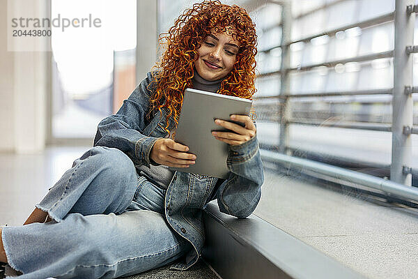 Smiling redhead young woman with curly hair using tablet PC sitting on floor