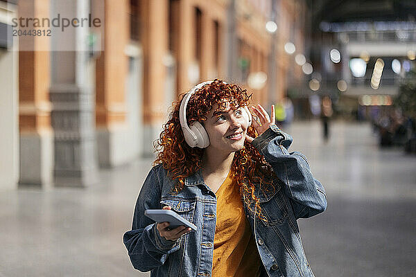 Smiling redhead young woman listening to music through wireless headphones walking with smart phone