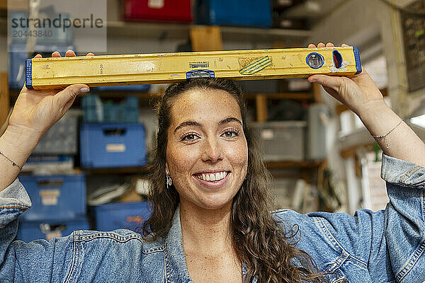 Smiling carpenter holding leveling tool on head at workshop