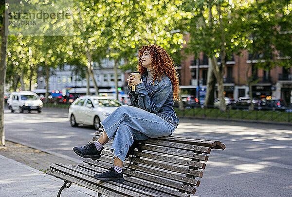 Young redhead woman sitting on bench with reusable coffee cup at sidewalk