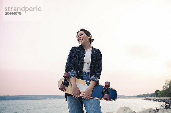 Young woman holding skateboard by sea at sunset