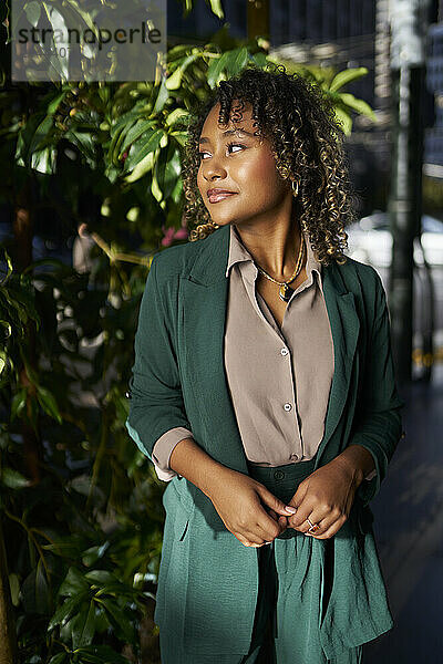 Smiling young businesswoman looking away while standing in front of plant