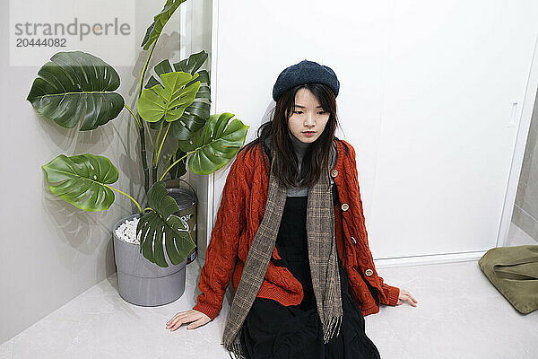 Young woman with warm clothes sitting by potted plant