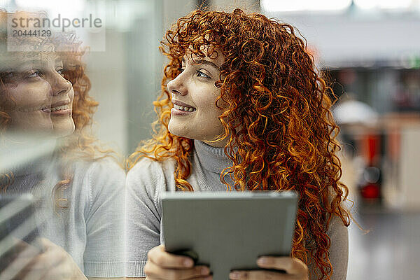 Smiling young redhead woman with tablet PC looking out through window