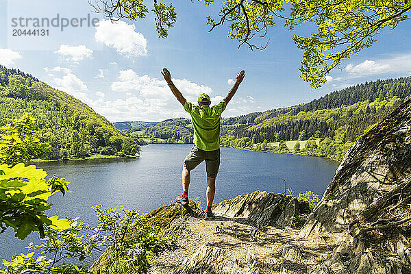 Man with arms raised standing on rock in front of Obersee lake at north Rhine Westphalia  Eifel national park  Germany