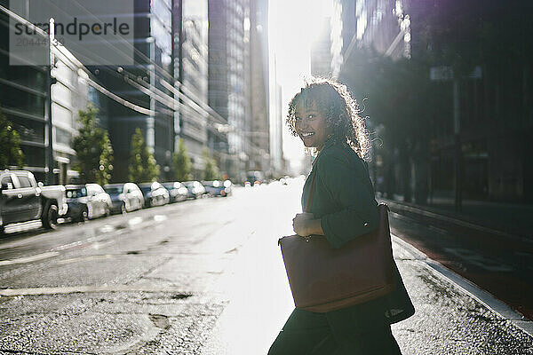 Smiling young businesswoman with purse crossing street in city