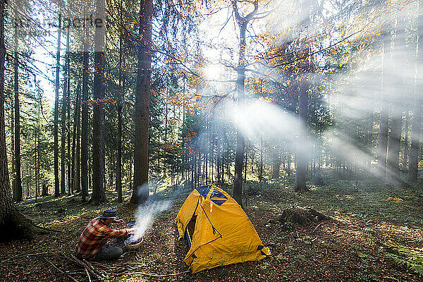 Senior man preparing food near tent and trees in forest