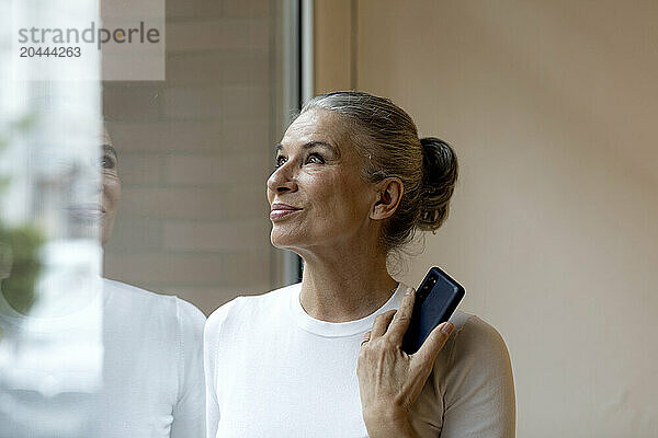 Smiling senior woman with smart phone looking out through window at home