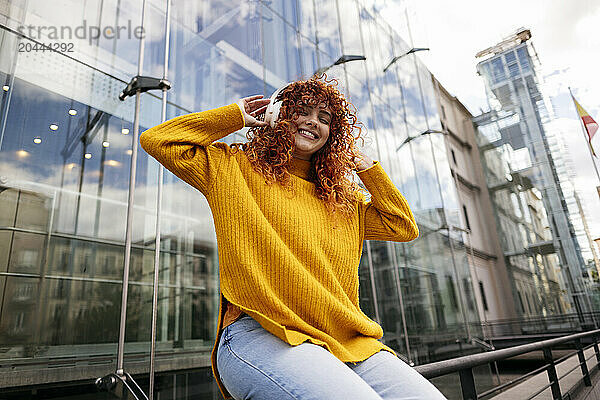 Happy redhead woman listening to music through wireless headphones in front of building