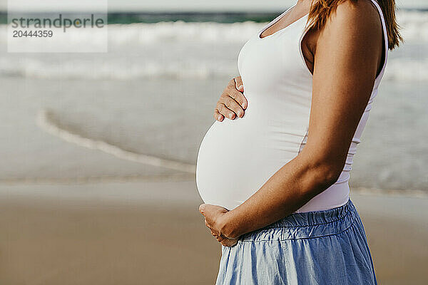 Pregnant woman standing with hands on stomach at beach