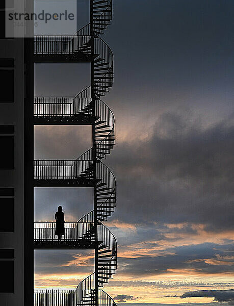 Silhouette of woman standing on fire escape looking at cloudy sky at dusk