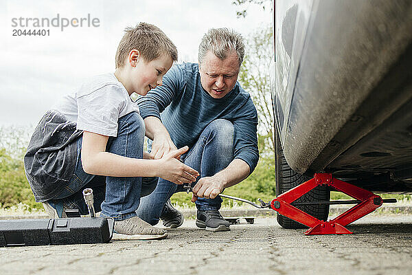 Son helping father installing car jack below car