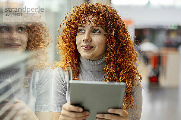 Smiling redhead young woman with tablet PC looking out through window