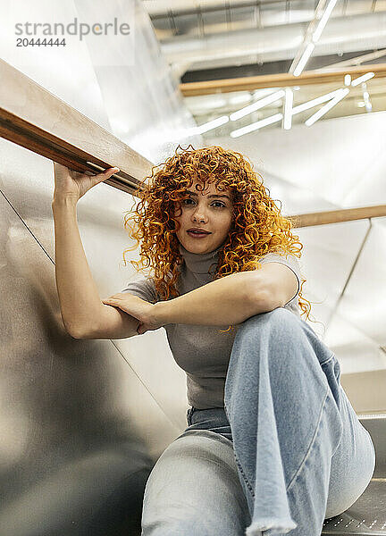 Confident beautiful redhead young woman with curly hair sitting on staircase