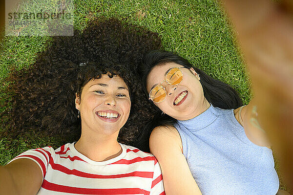 Laughing friends taking selfie and lying on grass at park