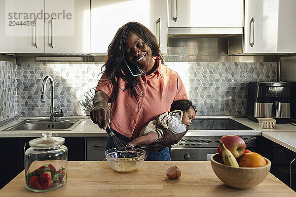 Mature woman talking on phone and holding baby girl in kitchen