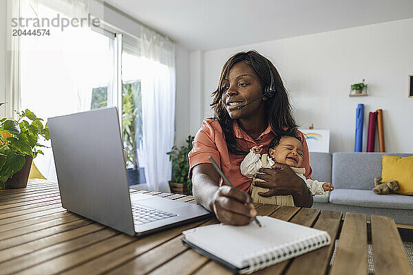 Working mother writing in diary and holding daughter at home office