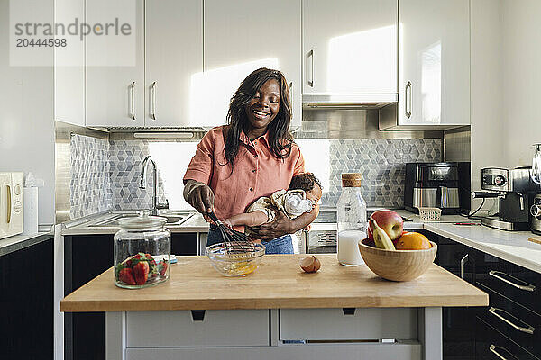 Happy mother preparing food and holding baby in kitchen at home