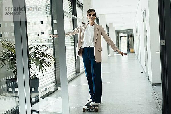 Businesswoman with arms outstretched skateboarding in office corridor