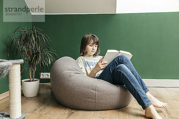 Teenage girl reading book on pouffe at home