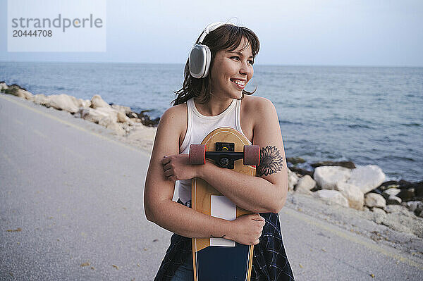 Smiling woman embracing skateboard listening to music through wireless headphones