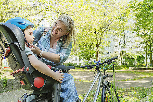 Mother fastening cycling helmet of son sitting on bicycle seat in park