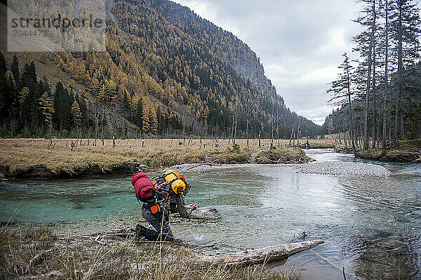 Senior man with backpack kneeling near river in front of mountains