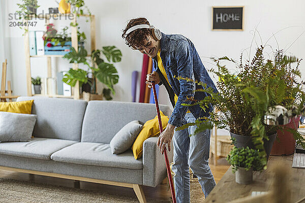 Happy man wearing wireless headphones and sweeping with broom at home