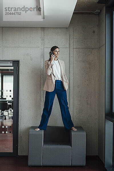 Businesswoman standing on armchair and talking on smart phone at office