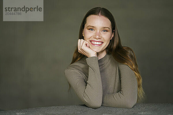 Happy woman with hand on chin sitting at table in front of gray wall