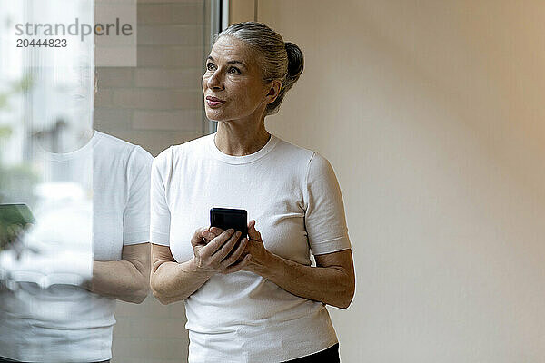 Senior woman with mobile phone leaning on window at home