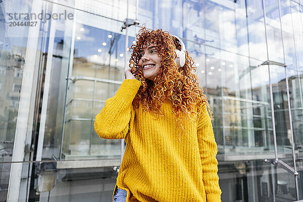 Happy redhead woman listening to music through wireless headphones in front of modern building