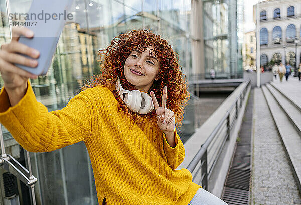 Beautiful young redhead woman showing peace sign taking selfie through mobile phone