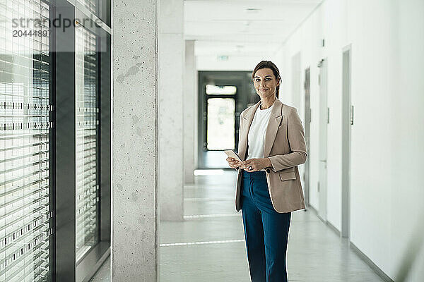 Cheerful businesswoman holding smart phone and standing at office