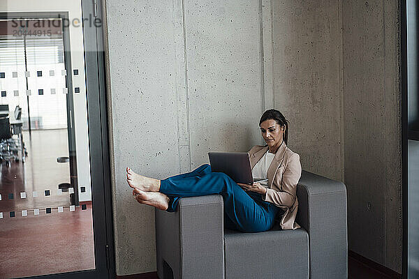 Businesswoman sitting on armchair with feet up and using laptop at office