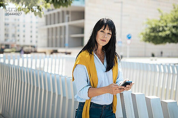 Confident businesswoman with smart phone leaning on railing