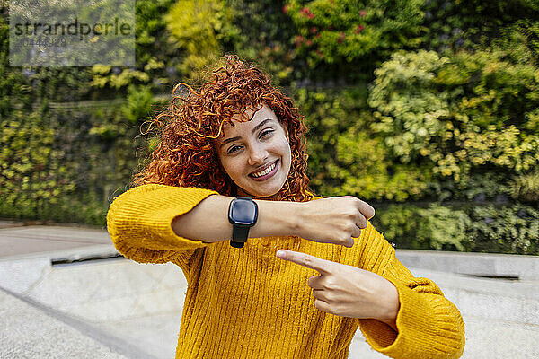 Happy redhead young woman with curly hair pointing at smart watch