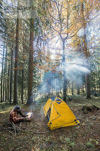 Senior man cooking food near yellow tent in forest