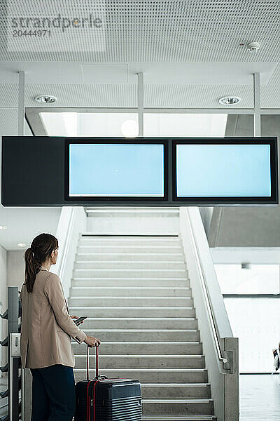 Businesswoman holding smart phone and suitcase at airport terminal