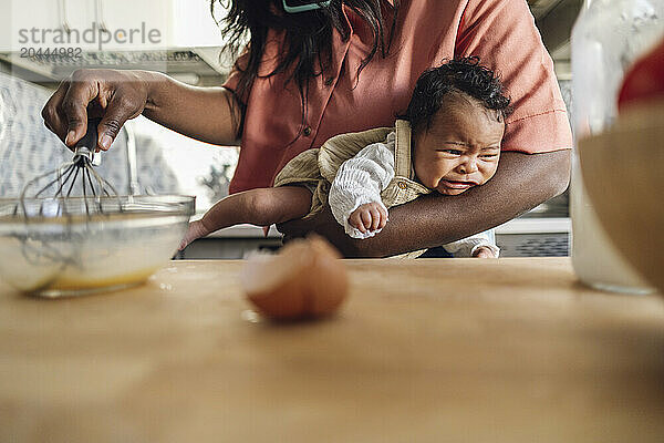 Mother whisking egg and holding crying daughter in kitchen