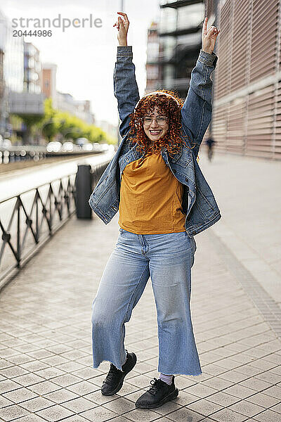 Happy redhead young woman listening to music through wireless headphones enjoying with arms raised in city