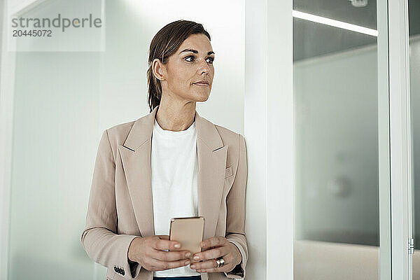 Business professional holding smart phone and standing near wall at office