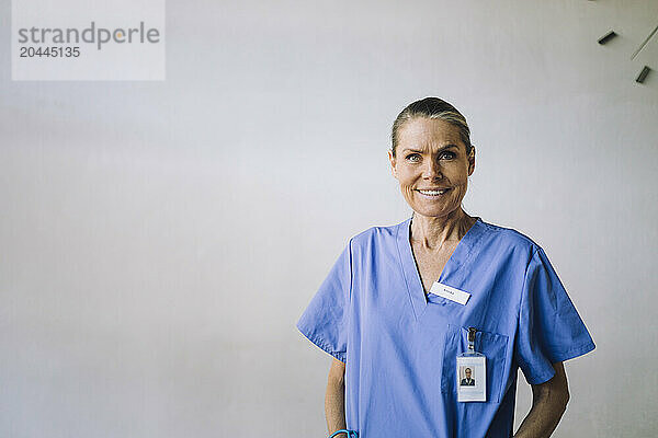 Portrait of smiling senior female doctor standing in front of white wall at hospital